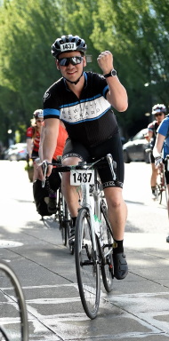 a man wearing cycling kit and sunglasses, raising a fist in triumph